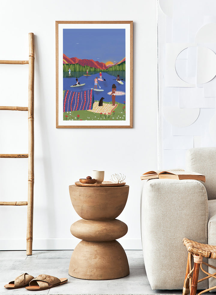 A framed art print featuring a colorful, stylized illustration of people paddleboarding and relaxing on a lake with mountains in the background. Paddleboard Lake Life by Ceyda Alasar outdoor art print featuring paddleboarders, mountain scenery, wildflowers, picnic blankets, and a lakeside dog.