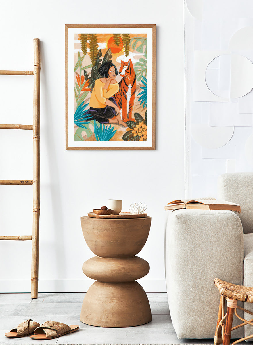 A framed art print featuring a stylized illustration of a woman in a yellow top and black skirt sitting beside an orange tiger, surrounded by tropical leaves and a warm orange sky. Woman with Tiger by Caroline Bonne Müller tropical art print featuring a woman beside a tiger among lush foliage, bold color, and hand-drawn texture.