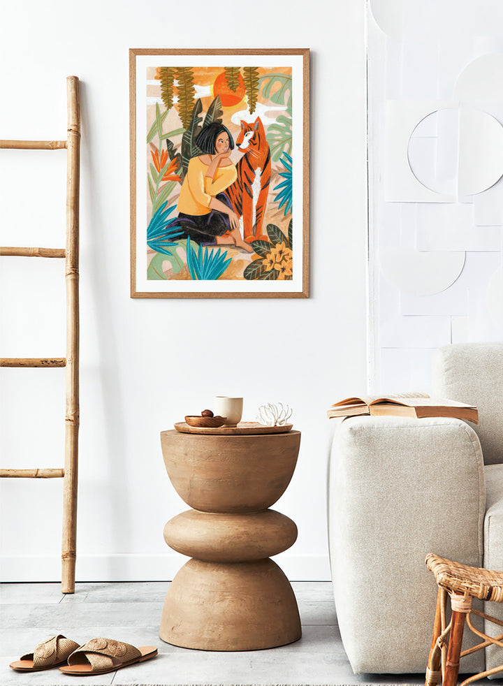 A framed art print featuring a stylized illustration of a woman in a yellow top and black skirt sitting beside an orange tiger, surrounded by tropical leaves and a warm orange sky. Woman with Tiger by Caroline Bonne Müller tropical art print featuring a woman beside a tiger among lush foliage, bold color, and hand-drawn texture.