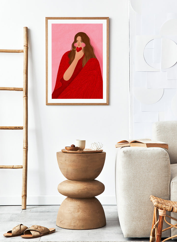 A framed art print featuring a stylized illustration of a woman with long brown hair, wearing a red patterned wrap, holding a red apple to her lips, set against a pink background. Cozy Woman Apple by Bea Müller portrait art print featuring a woman eating a red apple in bold red and pink tones for colorful modern wall decor.