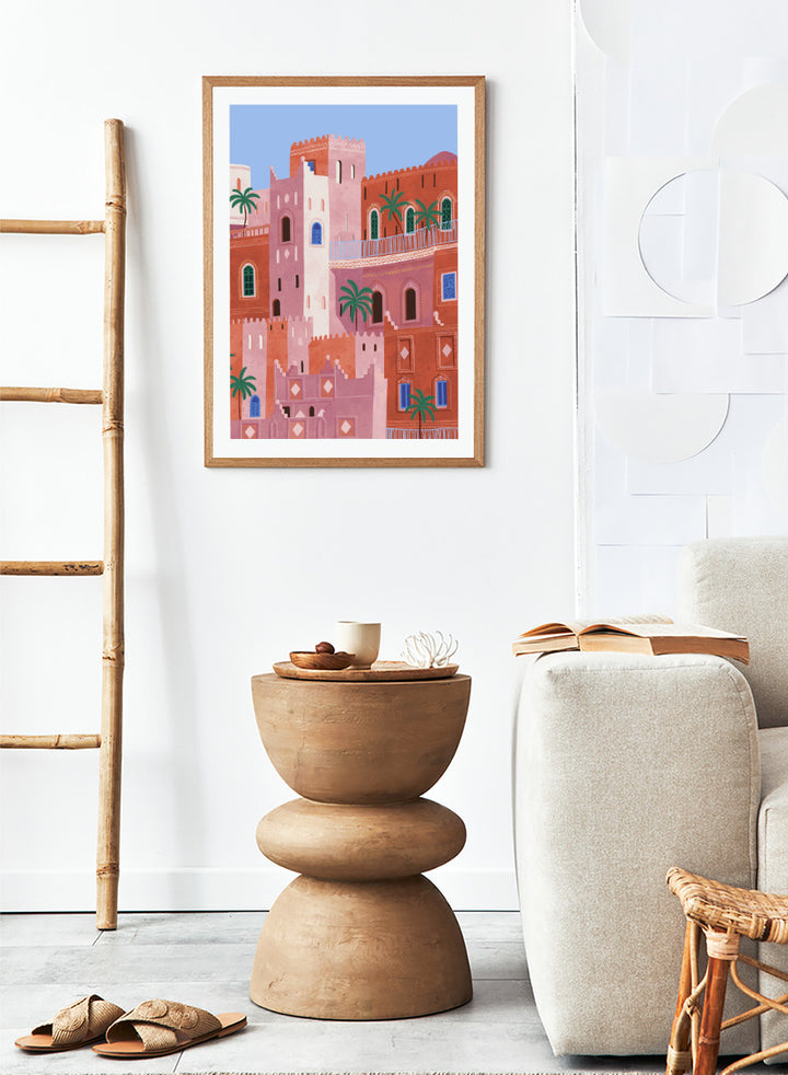 A framed art print featuring a stylized illustration of Moroccan-style buildings in shades of terracotta, pink, and cream, accented with green palm trees and blue window details, set against a light blue sky. Vibrant Moroccan City by Ceyda Alasar travel art print featuring layered terracotta architecture, arched windows, palms, and colorful Moroccan details.