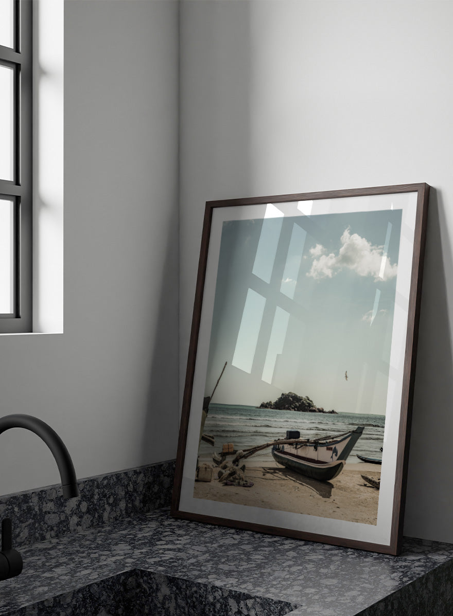 A framed photographic print of a coastal beach scene with a small boat on the sandy shore, the sea and distant rocky island, and a pale blue sky with clouds. Seaside Serenity by Shot by Clint coastal photography print featuring a fishing boat, sandy shore, ocean waves, distant island, and weathered seaside details.