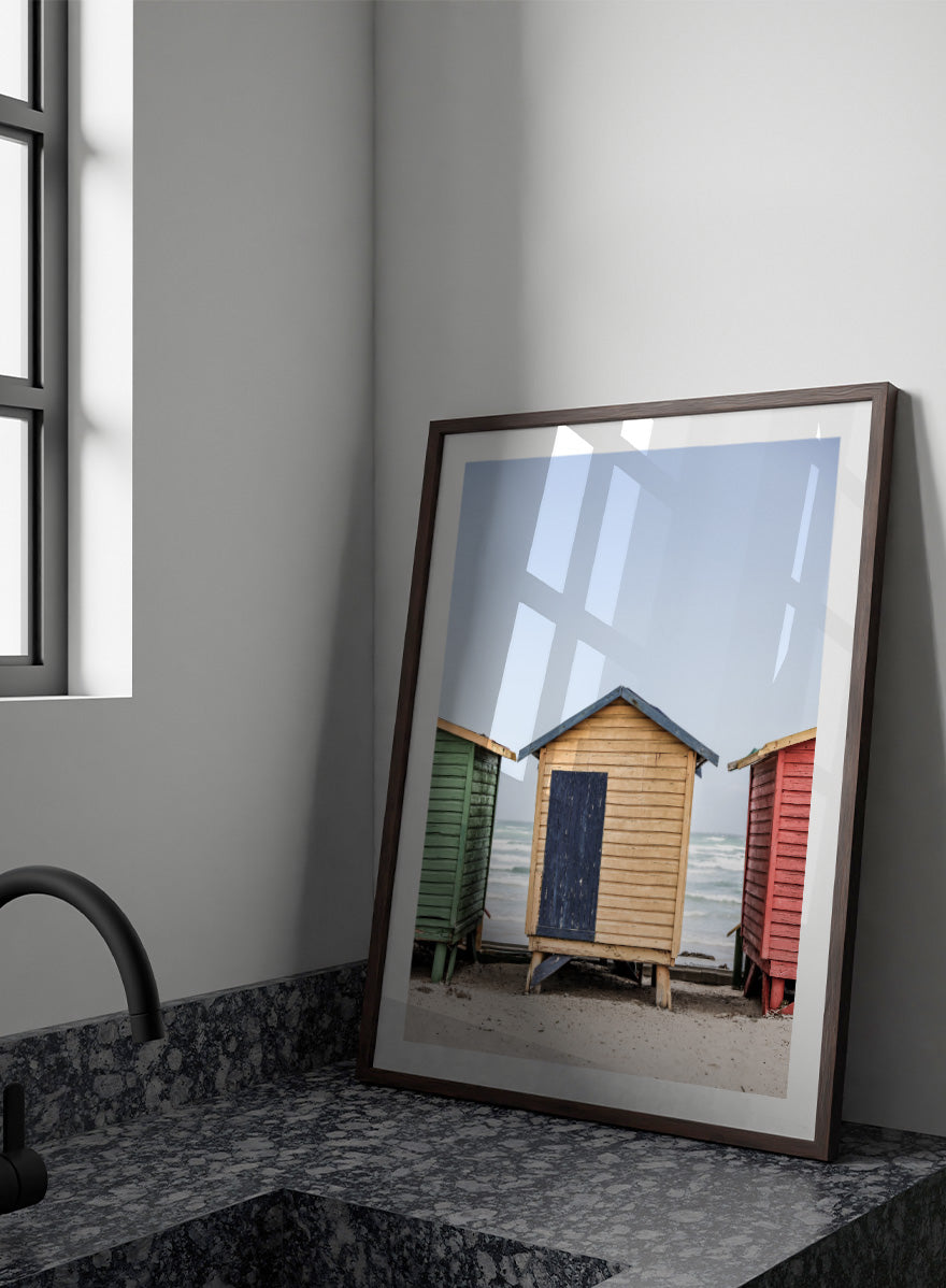 A framed photographic print of three colorful wooden beach huts on a sandy beach, with the central hut in natural wood and a dark blue door, flanked by green and red huts under a pale blue sky. Colorful Beach Huts by Shot by Clint coastal photography print featuring green, natural wood, and red huts with weathered texture and ocean views.