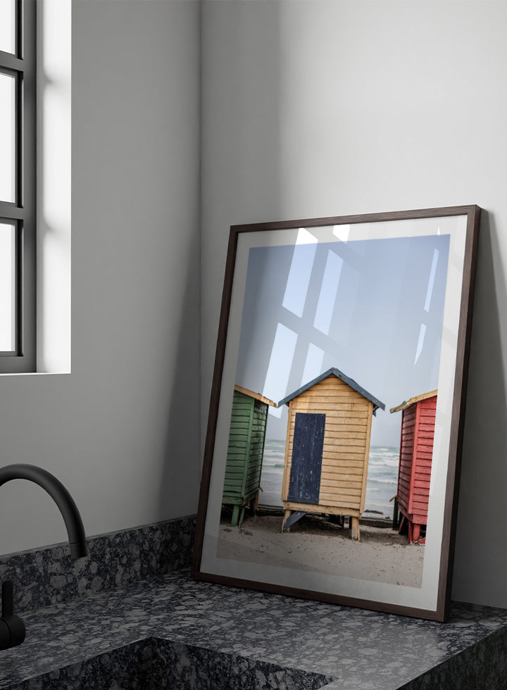 A framed photographic print of three colorful wooden beach huts on a sandy beach, with the central hut in natural wood and a dark blue door, flanked by green and red huts under a pale blue sky. Colorful Beach Huts by Shot by Clint coastal photography print featuring green, natural wood, and red huts with weathered texture and ocean views.