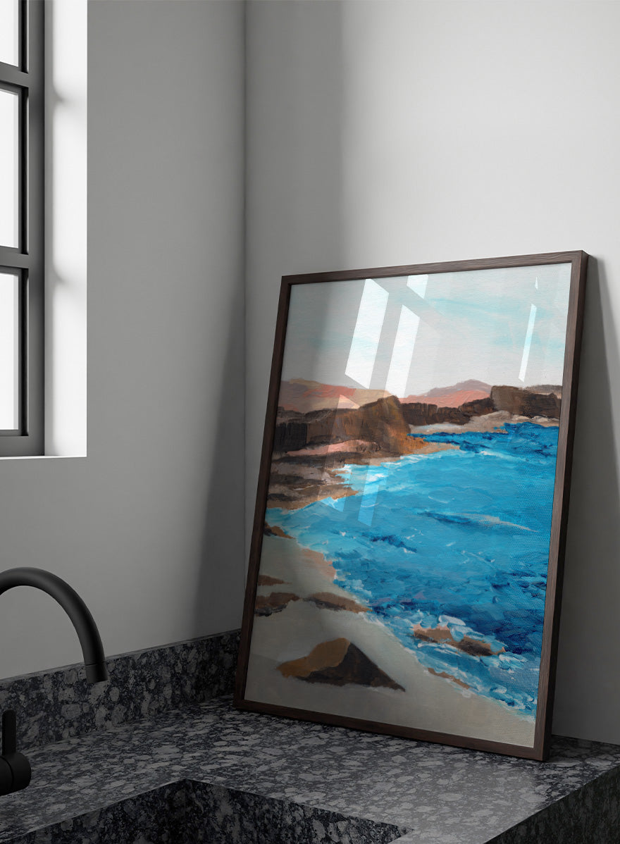 A framed landscape painting depicting a coastal scene with bright blue ocean water, sandy shore, dark rocky cliffs, and distant hills under a pale sky. Shop Rocky Coastal Waters by Nikita Jariwala, a coastal wall art print poster with blue ocean water, rocky cliffs, and a peaceful seaside view.