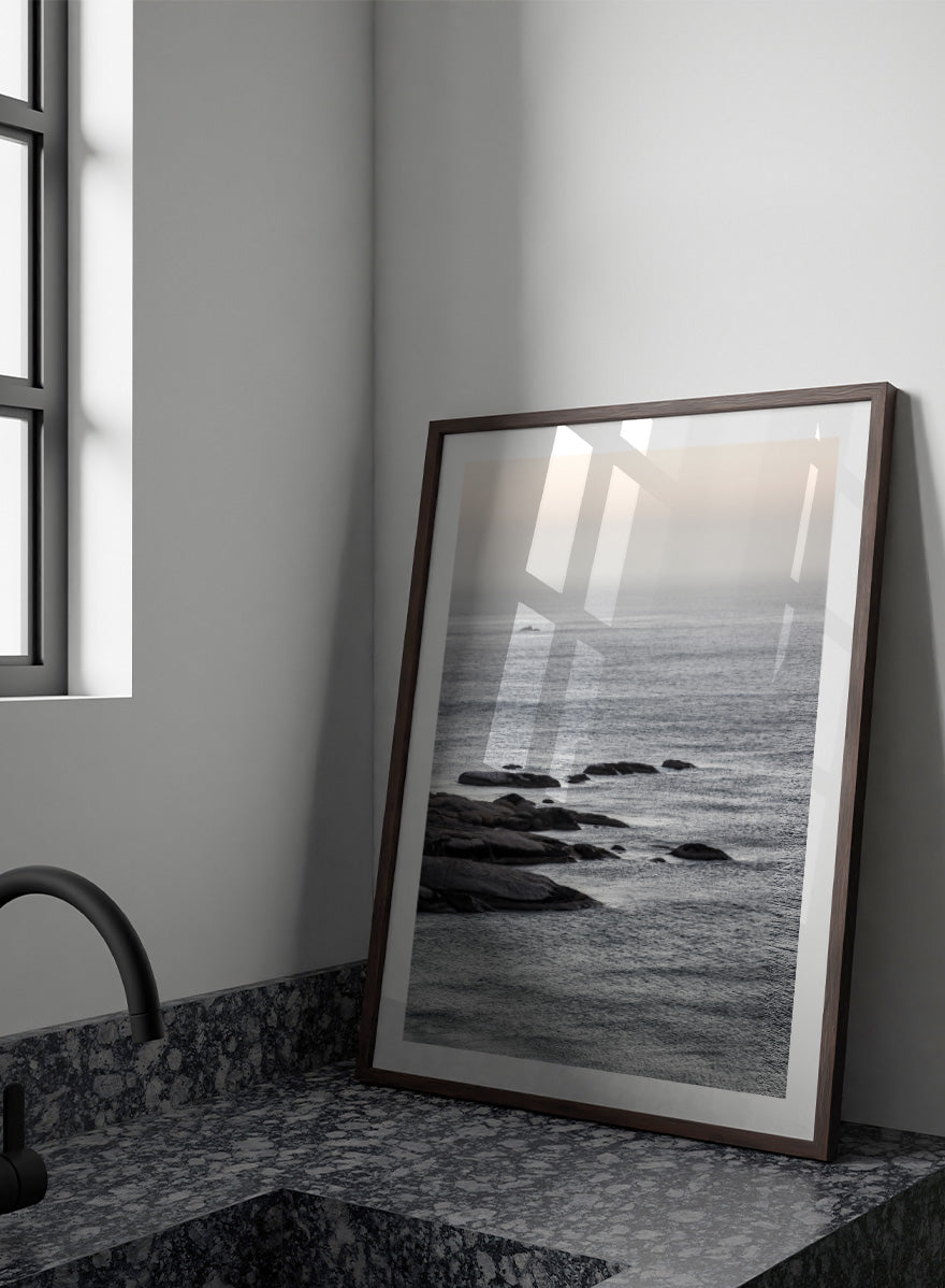 A framed black and white coastal landscape photograph featuring a calm sea, distant rocks, and a pale sky. Silver Sands by Shot by Clint coastal photography print featuring silver-blue water, dark shoreline rocks, and a soft misty horizon.