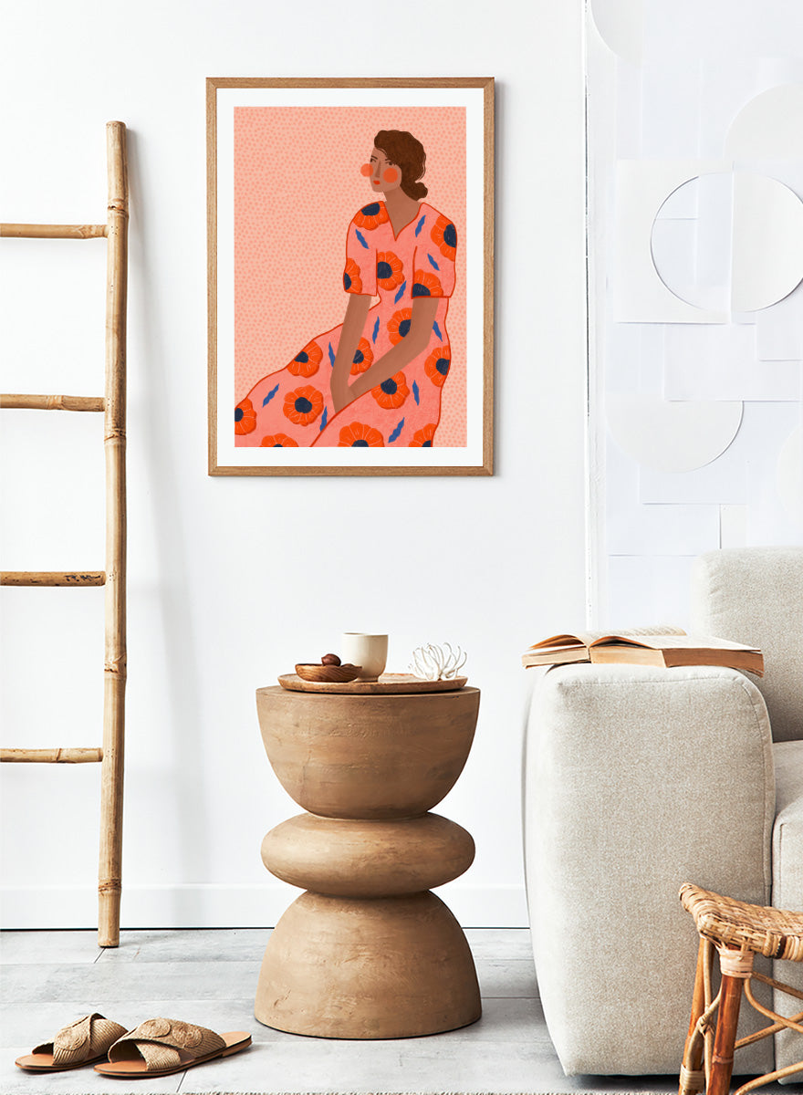 A stylized fashion illustration of a seated woman in a coral pink dress with large orange poppy motifs and blue leaf accents, shown with a white border and black frame. Stylish Poppy Portrait by Bea Müller floral woman art print featuring a pink dress covered in orange poppies and blue leaves for colorful modern wall decor.