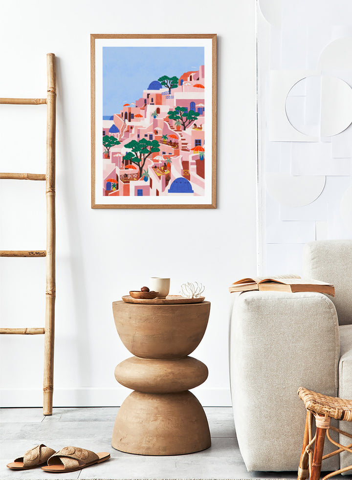 A stylized art print depicting a hillside Mediterranean village with pink and peach buildings, blue domes, green trees, and a light blue sky. Santorini Summer Dream by Ceyda Alasar art print featuring blue domes, terraced Mediterranean architecture, orange umbrellas, and sunny Greek island charm.