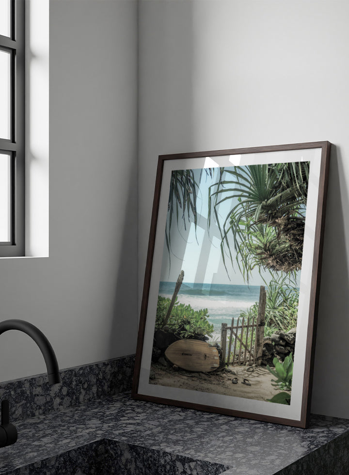 A framed beach scene art print featuring a wooden surfboard leaning against a rustic gate, with palm leaves framing a view of the ocean under a light blue sky. Coastal Serenity by Shot by Clint coastal art print featuring a surfboard, tropical plants, rustic gate, sandy path, and ocean waves.