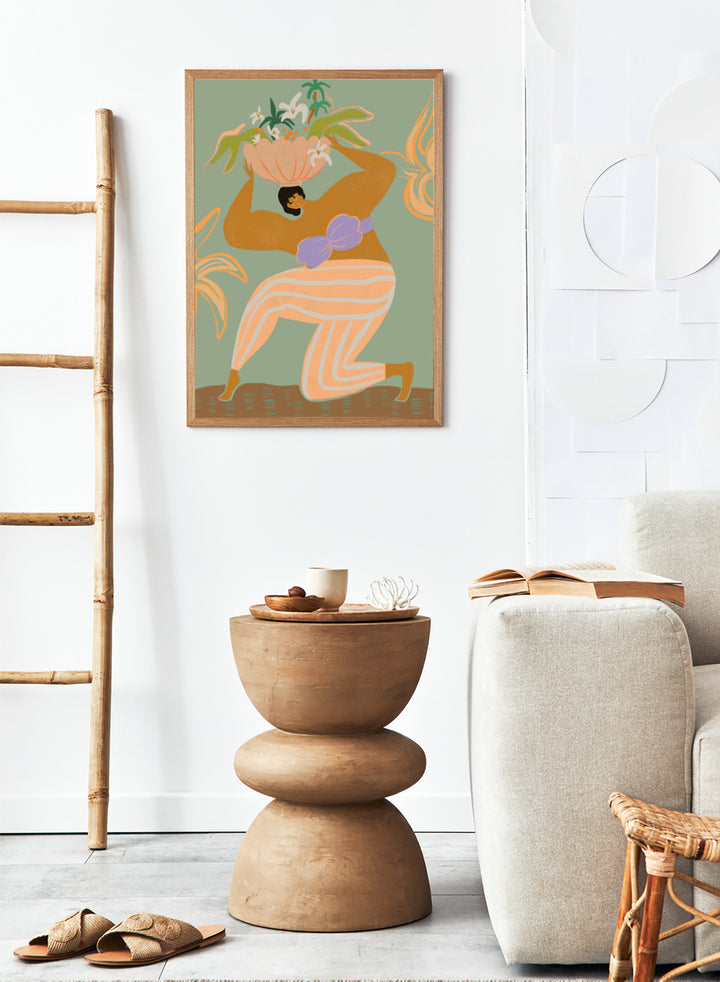 A stylized art print featuring a woman in flowing peach and white striped pants with a purple sash, carrying a large flower basket on her head against a muted sage green background. The Weight of the World by Arty Guava premium framed fine art print featuring a woman carrying tropical flowers in soft green, peach, and lilac tones.