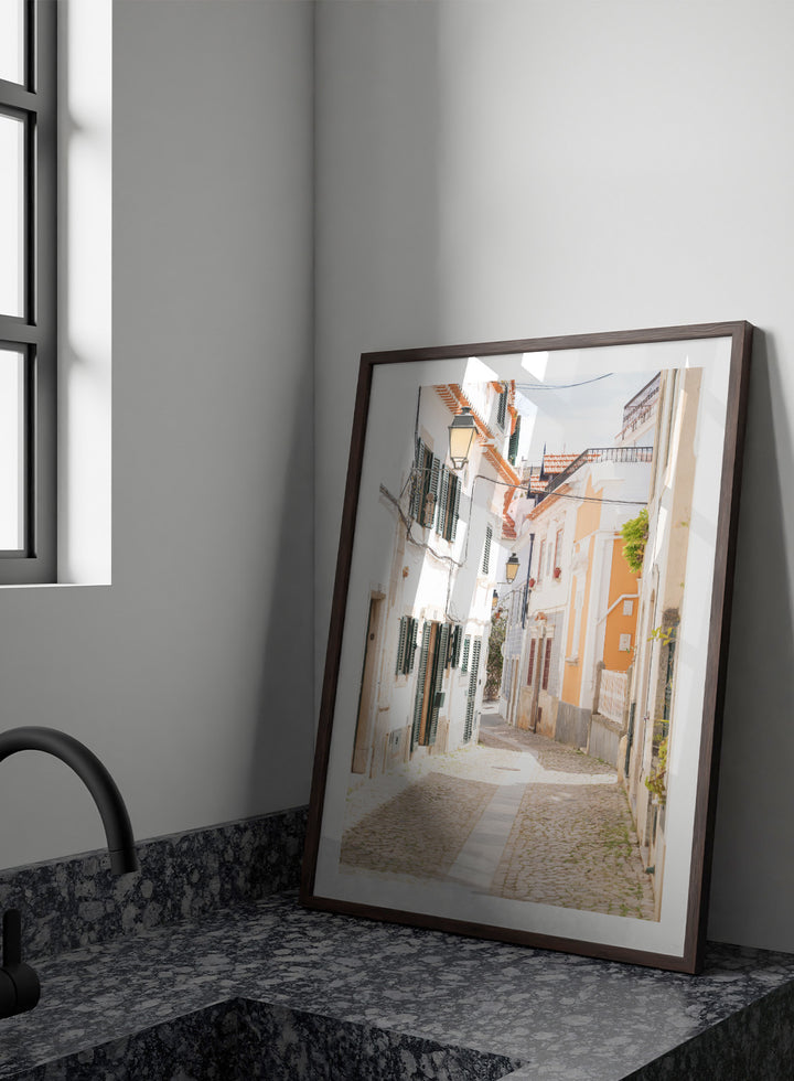 Cobbled street in a quaint European town with white buildings and a clear sky. Charming European Cobblestone Street by Henrike Schenk is a travel photography print featuring cobblestone streets, historic architecture, and European wall art.