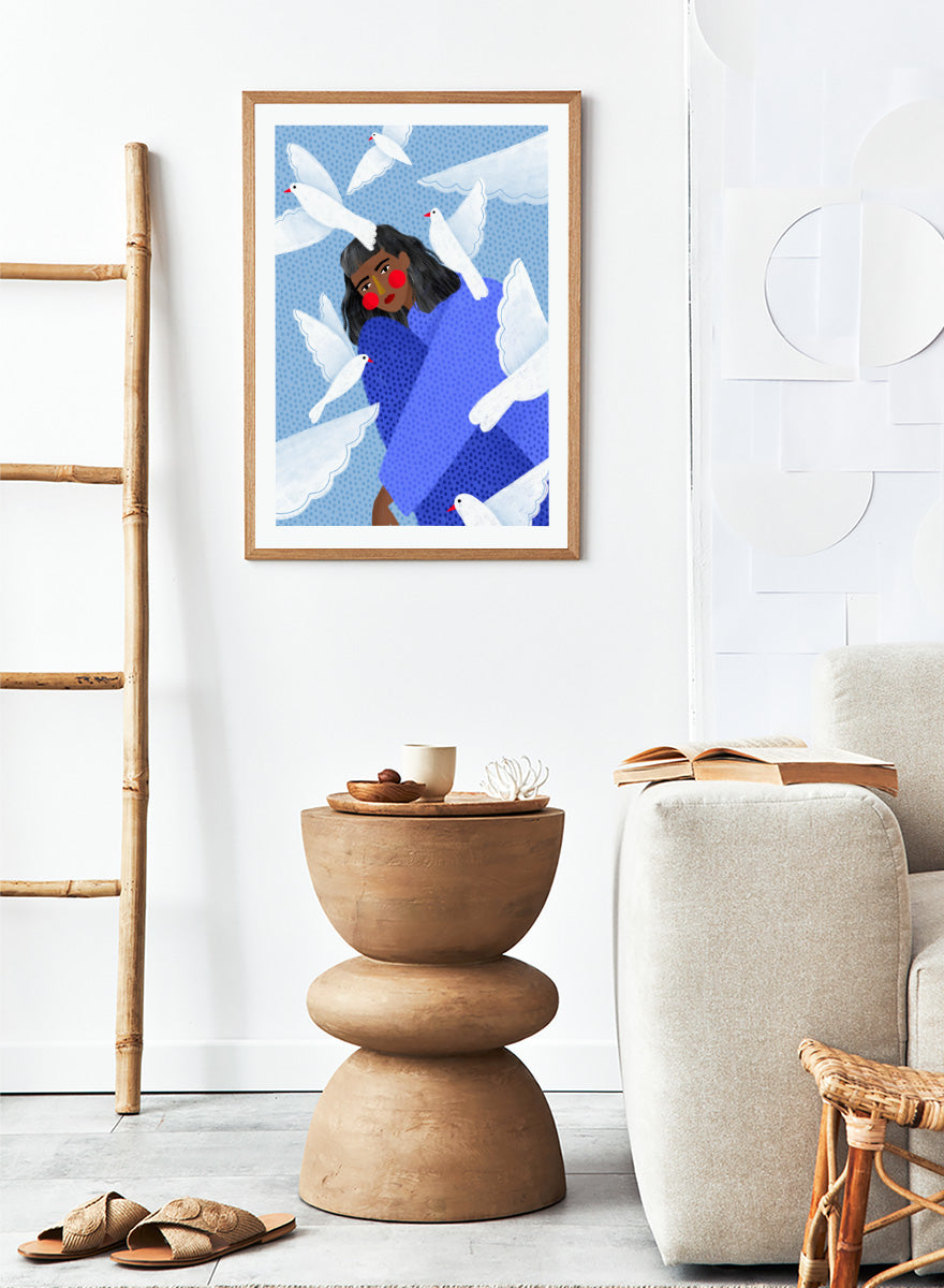 A framed art print featuring a stylized illustration of a woman with dark hair and red cheeks, wrapped in a blue patterned blanket, surrounded by white doves against a light blue dotted background. Peaceful Woman and Doves by Bea Müller art print featuring a woman in blue surrounded by white birds, layered textures, and soft modern color.