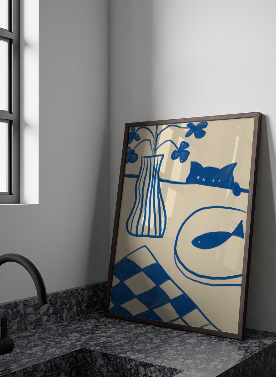 A framed art print featuring a hand-drawn style illustration in blue ink on a warm cream background. The design includes a striped vase with flowers, a cat peeking over a line, a fish on a plate, and a harlequin check pattern in the lower left corner. Blue Cat And Fish by The Art Concept is a playful cat art print featuring a curious blue cat, fish plate, striped vase, flowers, and checkered tablecloth.