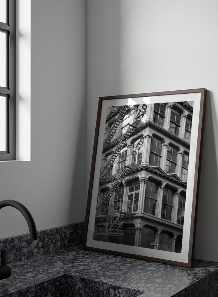 Black and white architectural photograph of a corner building facade with ornate windows and external fire escapes. Urban Geometry by Shot by Clint black-and-white architectural photography print featuring fire escapes, tall windows, ornate columns, and layered city detail.