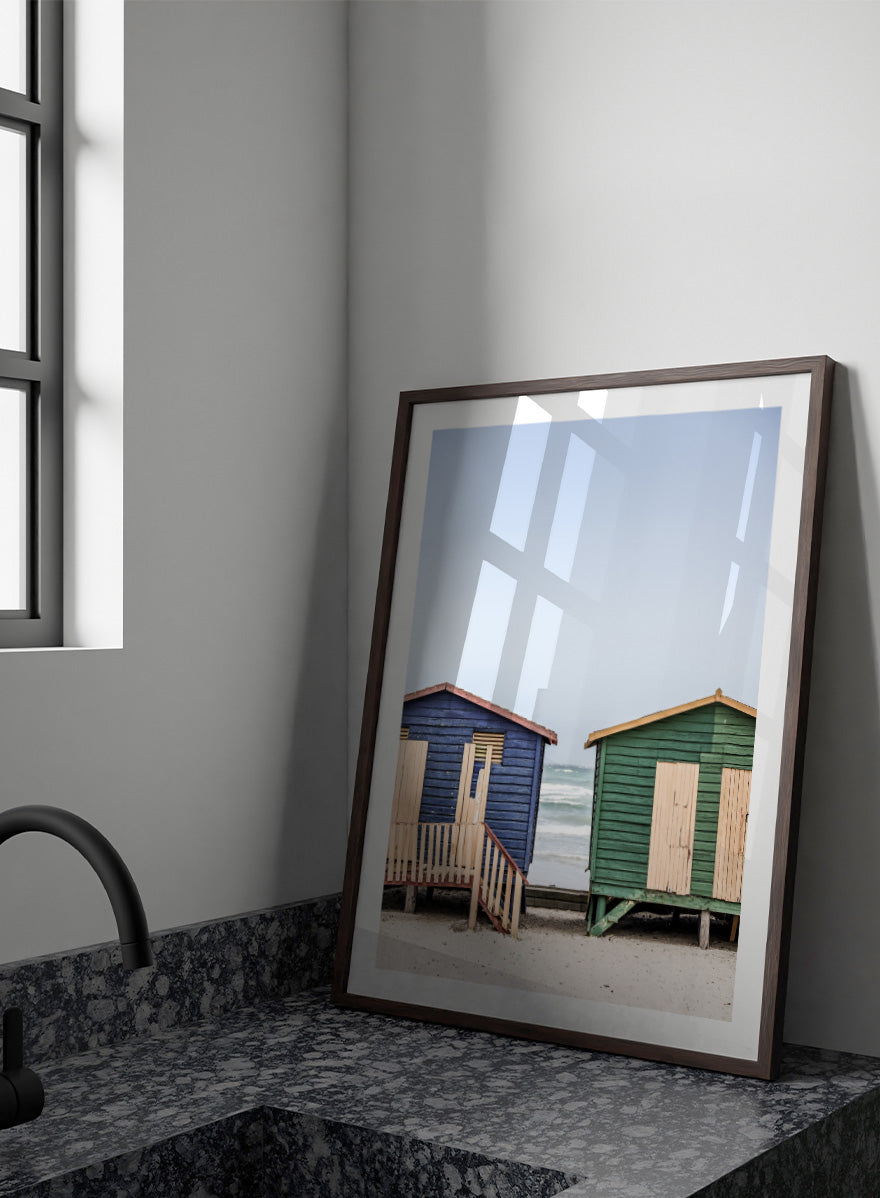 A framed photographic print of two wooden beach huts on a sandy beach under a pale blue sky, with one hut in dark blue and the other in green, both with light wood shutters and doors. Coastal Huts Harmony by Shot by Clint coastal photography print featuring blue and green beach huts, sandy ground, weathered wood, and ocean views.