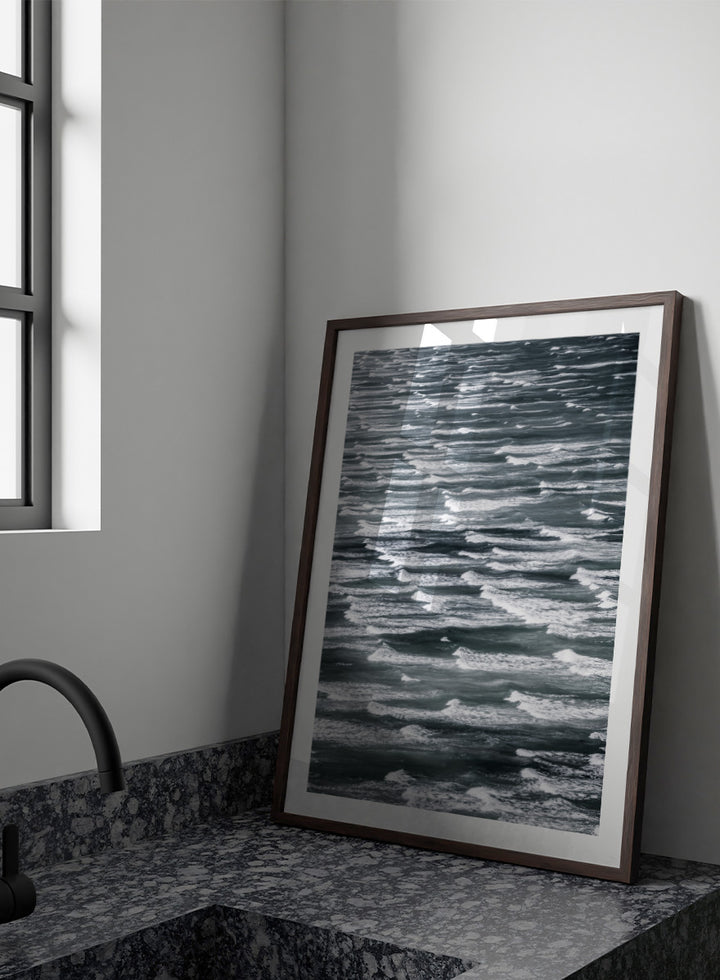 A framed black and white photographic print depicting a close-up view of ocean waves with white foam crests against dark water. Waves in Rhythm by Shot by Clint ocean photography print featuring layered blue-grey waves, white foam, and powerful natural movement across the sea.