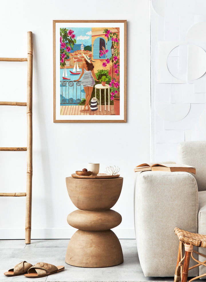 Woman on a balcony overlooking a coastal town with boats and flowers. Woman Balcony Coastal View by Caroline Bonne Müller is a fine art print and wall poster of a woman overlooking a coastal town with boats in a modern aesthetic style.