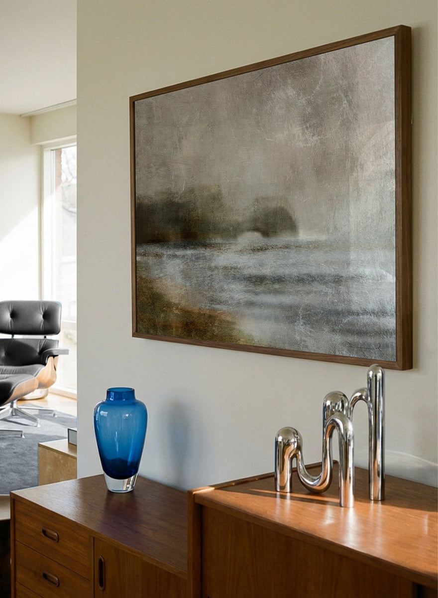A framed abstract landscape art print featuring textured waves and a distant rocky shoreline in muted beige, gray, brown, and white tones. Shop Dusk by Dan Hobday, a moody landscape art print featuring misty water, low hills, earthy neutral toned nature, and soft layered brush texture.