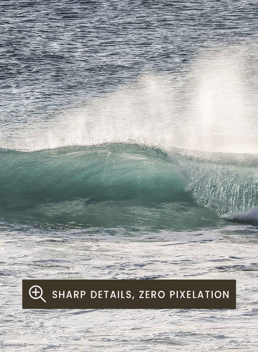 A framed photographic art print of an ocean scene, featuring a gentle wave breaking on a sandy beach under a pale sky. Tranquil Ocean Scene by Shot by Clint ocean photography print with a rolling wave, soft sea spray, and airy blue coastal tones.