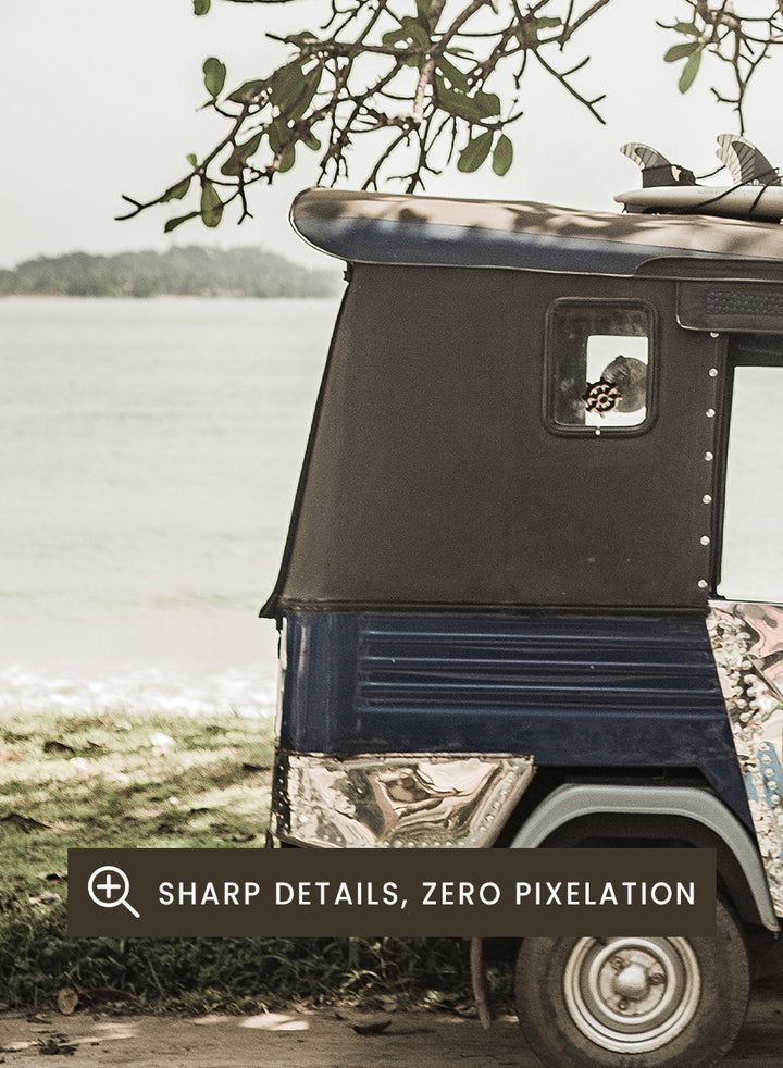 A framed photographic print featuring a small blue and white auto rickshaw parked under a large leafy tree beside a body of water. Tuk Tuk Tour by Shot by Clint travel photography print featuring a blue tuk tuk, surfboard, coastal tree, and relaxed seaside setting.