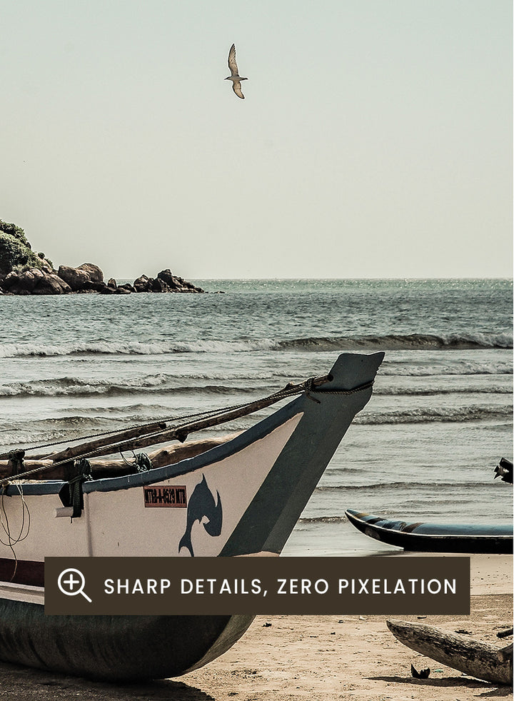 A framed photographic print of a coastal beach scene with a small boat on the sandy shore, the sea and distant rocky island, and a pale blue sky with clouds. Seaside Serenity by Shot by Clint coastal photography print featuring a fishing boat, sandy shore, ocean waves, distant island, and weathered seaside details.