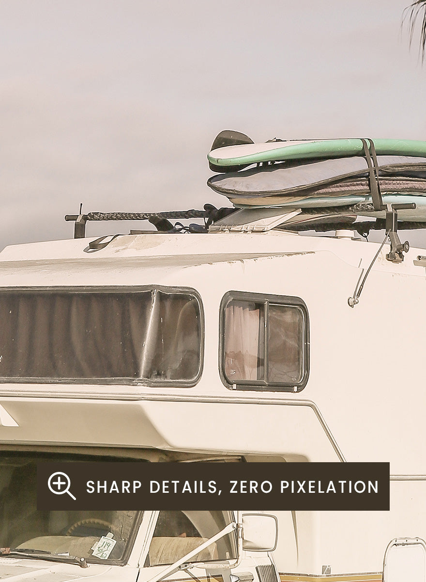 A vintage white camper truck parked outdoors near palm trees and a fence, with a surfboard strapped to its roof rack. Vintage Camper Parked by Shot by Clint travel photography print featuring a retro camper, surfboards, palm trees, and sun-faded coastal tones.