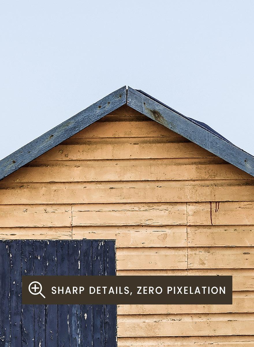 A framed photographic print of three colorful wooden beach huts on a sandy beach, with the central hut in natural wood and a dark blue door, flanked by green and red huts under a pale blue sky. Colorful Beach Huts by Shot by Clint coastal photography print featuring green, natural wood, and red huts with weathered texture and ocean views.