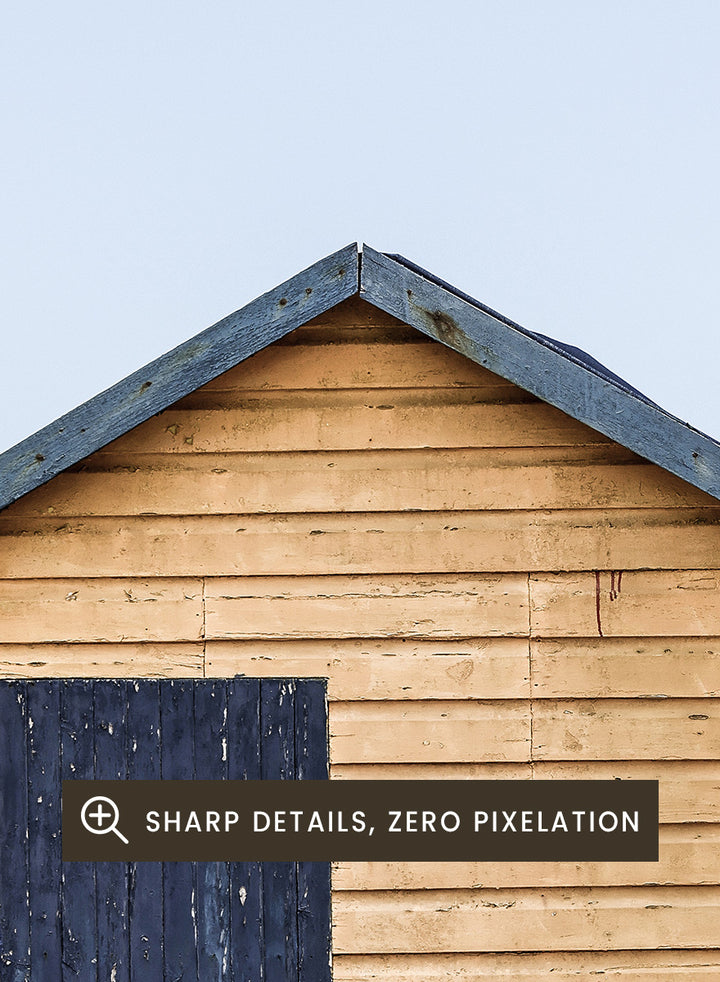 A framed photographic print of three colorful wooden beach huts on a sandy beach, with the central hut in natural wood and a dark blue door, flanked by green and red huts under a pale blue sky. Colorful Beach Huts by Shot by Clint coastal photography print featuring green, natural wood, and red huts with weathered texture and ocean views.