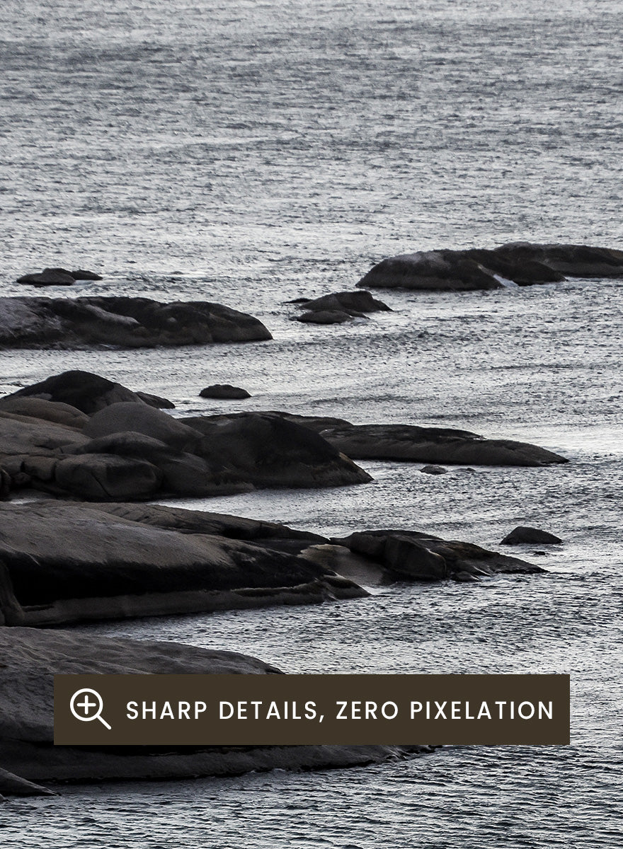 A framed black and white coastal landscape photograph featuring a calm sea, distant rocks, and a pale sky. Silver Sands by Shot by Clint coastal photography print featuring silver-blue water, dark shoreline rocks, and a soft misty horizon.