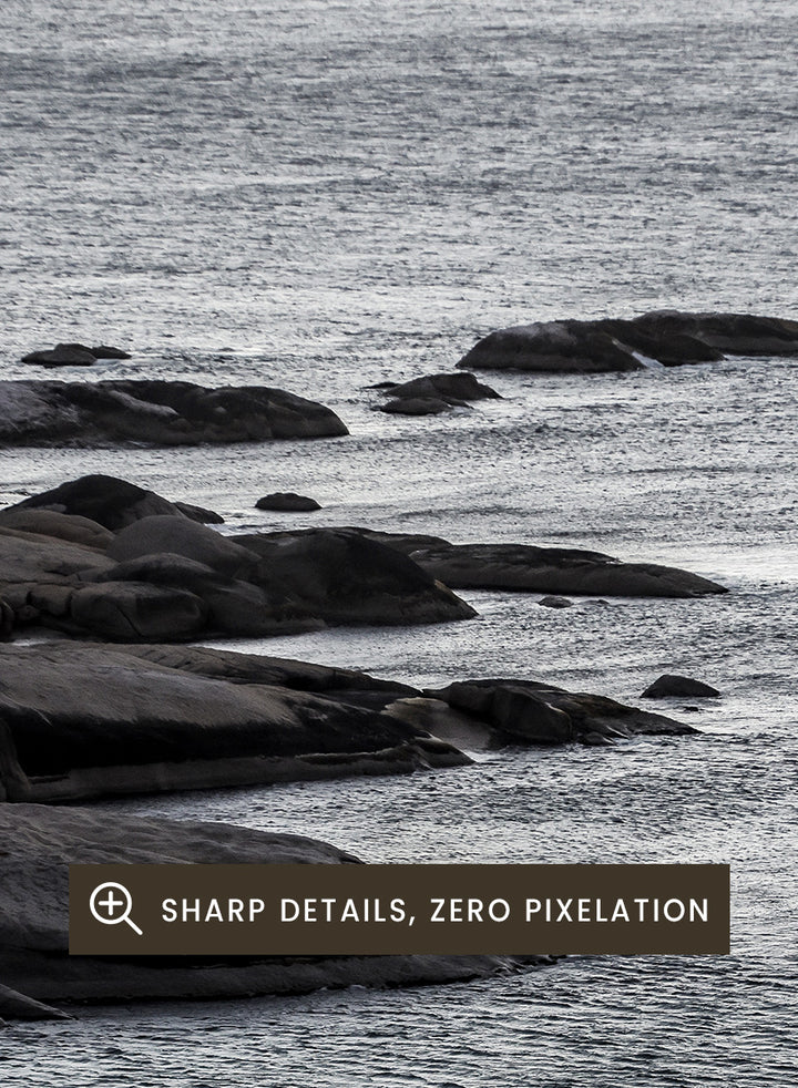 A framed black and white coastal landscape photograph featuring a calm sea, distant rocks, and a pale sky. Silver Sands by Shot by Clint coastal photography print featuring silver-blue water, dark shoreline rocks, and a soft misty horizon.