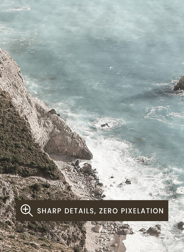 A framed photographic art print depicting a coastal landscape with a steep rocky cliff overlooking a calm, pale blue sea with a small rock formation in the foreground. Coastal Serenity by Shot by Clint coastal photography print featuring pale blue water, rugged cliffs, rocky shores, and a soft hazy horizon.