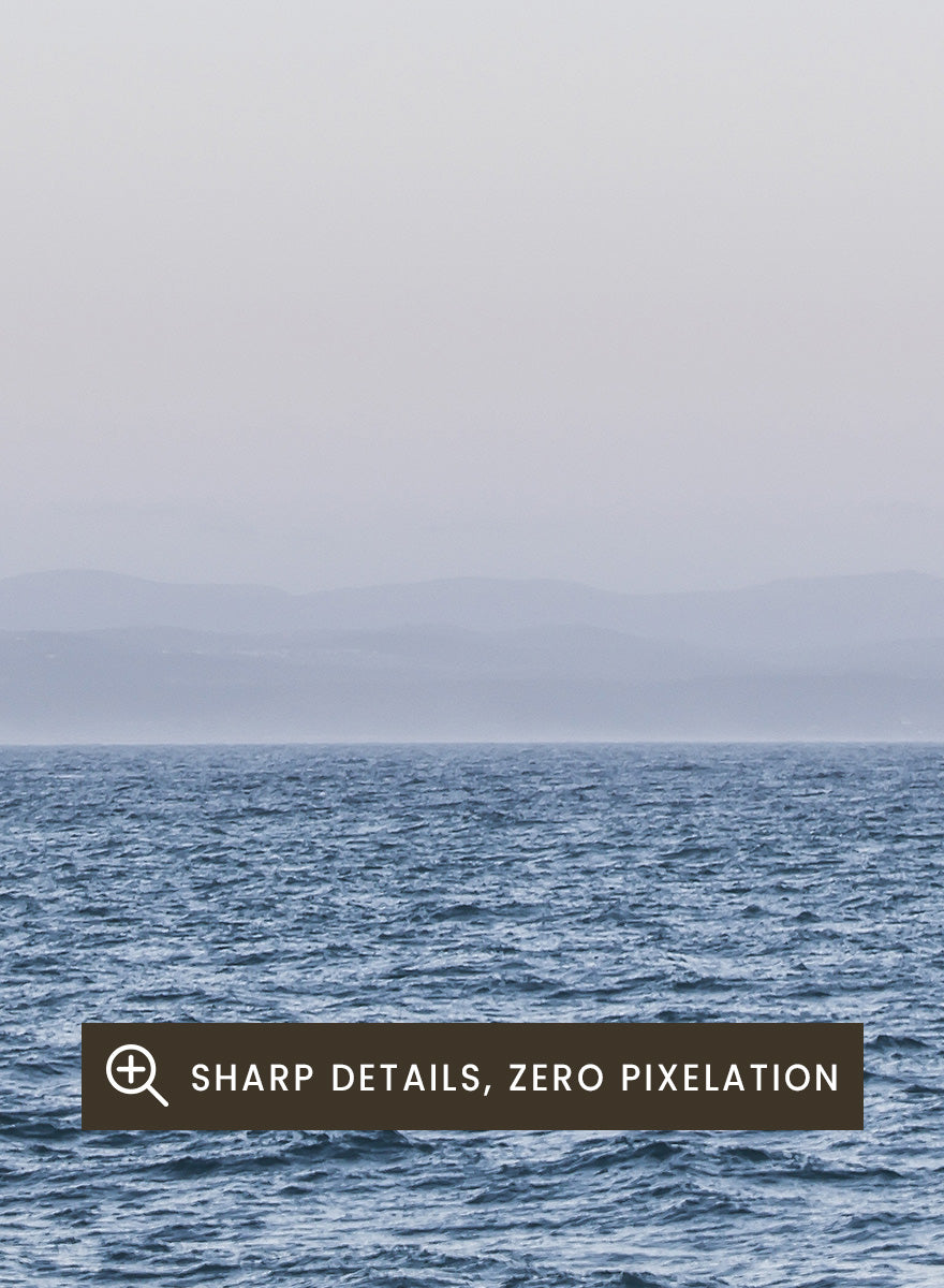 A minimalist framed art print depicting a calm blue ocean under a pale sky with a small visible moon. Moonlit Horizon by Shot by Clint coastal photography print featuring a pale moon, distant shoreline, open blue water, and soft lavender evening tones.