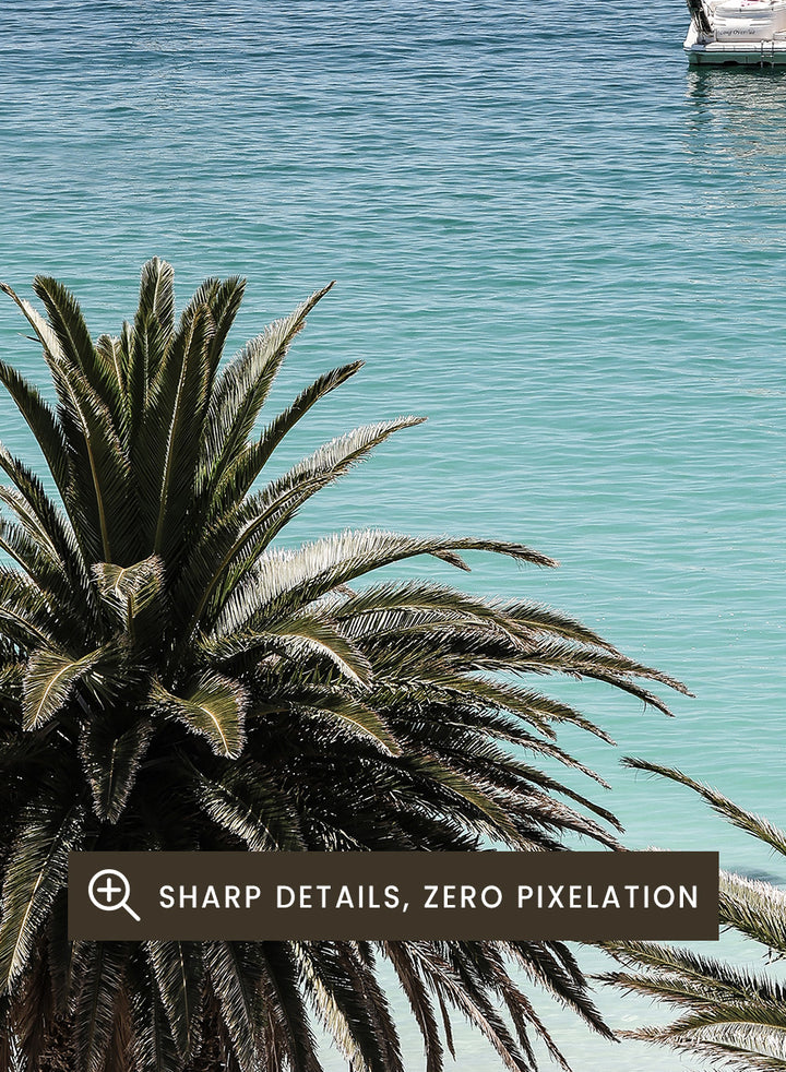 A framed coastal photograph showing a calm blue ocean with three boats, turquoise water near a white sandy shore, and two palm trees in the foreground. Tropical Serenity by Shot by Clint coastal photography print featuring turquoise water, sailboats, palm trees, and a bright tropical shoreline.
