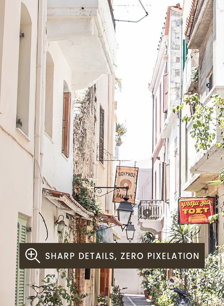  narrow street with a bakery sign in a European town. Charming Bakery Street by Henrike Schenk is a Greek street photography print featuring a Mediterranean bakery alley, pastel buildings, café seating, and travel wall art.