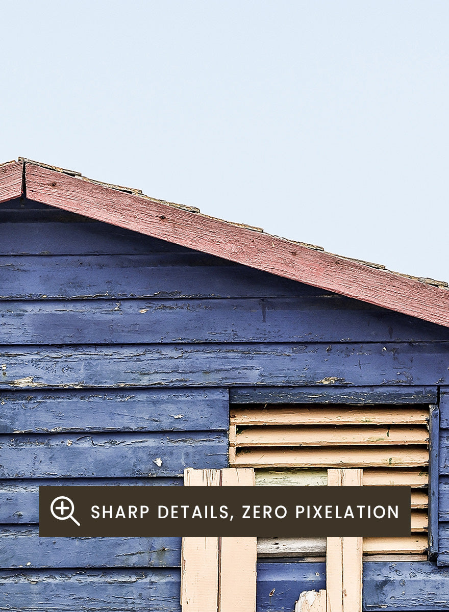 A framed photographic print of a blue wooden beach lifeguard hut on a sandy shore with the ocean and pale sky in the background. Beachside Retreat by Shot by Clint features a weathered blue beach hut and soft ocean backdrop in a calm coastal photography print.