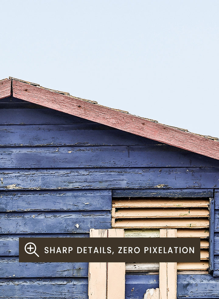 A framed photographic print of a blue wooden beach lifeguard hut on a sandy shore with the ocean and pale sky in the background. Beachside Retreat by Shot by Clint features a weathered blue beach hut and soft ocean backdrop in a calm coastal photography print.