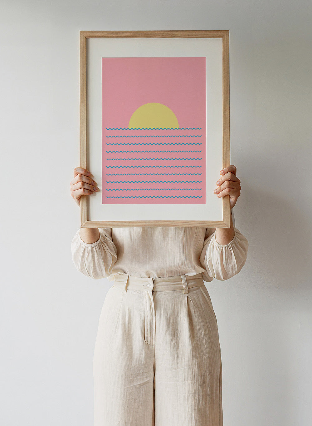 Simplified illustration of a sunset over water with blue waves on a pink background. Every Day the Sun Rises by Rosi Feist is a minimalist sun and waves art print with pink sky. Officially licensed modern poster with framing.