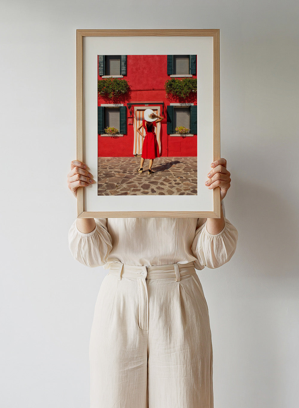 Woman in a red dress and white hat standing in front of a red wall with green shutters. Red Art Print by Anette Ohlendorf featuring a woman in a red dress standing against a red building. Officially licensed travel photography with strong color and symmetry.