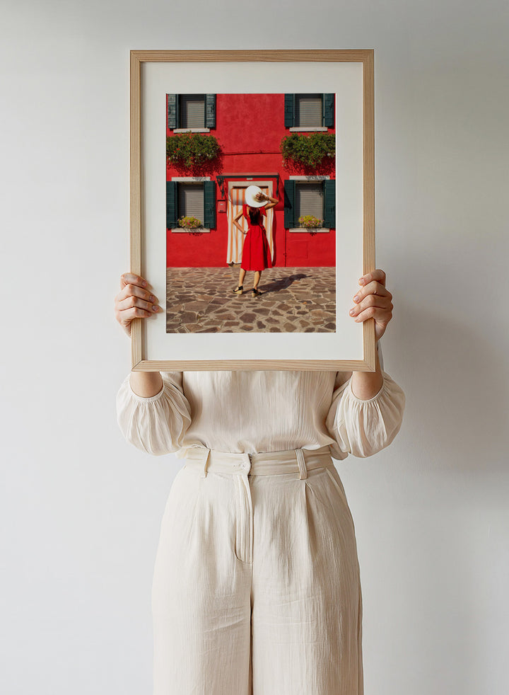 Woman in a red dress and white hat standing in front of a red wall with green shutters. Red Art Print by Anette Ohlendorf featuring a woman in a red dress standing against a red building. Officially licensed travel photography with strong color and symmetry.