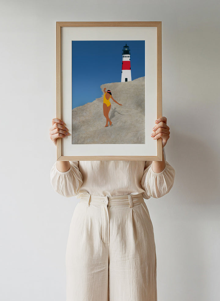 Woman in a yellow swimsuit standing on a rocky outcrop with a lighthouse in the background. My Beach by Jota de Jai is a modern coastal wall art print featuring a woman in a yellow swimsuit and a lighthouse. Officially licensed fine art poster with optional wood frame.
