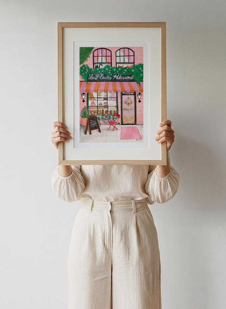Illustration of a bakery named 'Les Fleuris Patisserie' with a pink exterior and greenery. Bake Shop Front by Baroo Bloom art print featuring a colorful bakery storefront illustration. Whimsical European street scene wall poster with pink tones and playful detail.