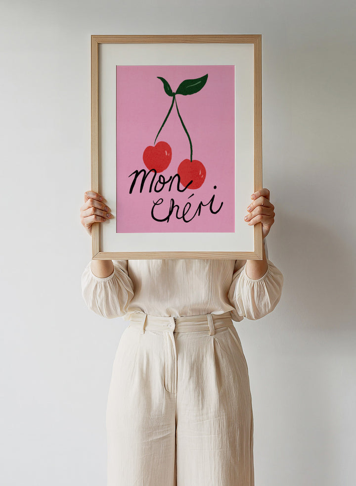 Two red cherries with green stems on a pink background and 'mon chéri' text. Mon Chéri by Dirkie Nell is a cherry typography art print featuring bold red fruit and playful lettering. A colorful love-inspired wall poster.