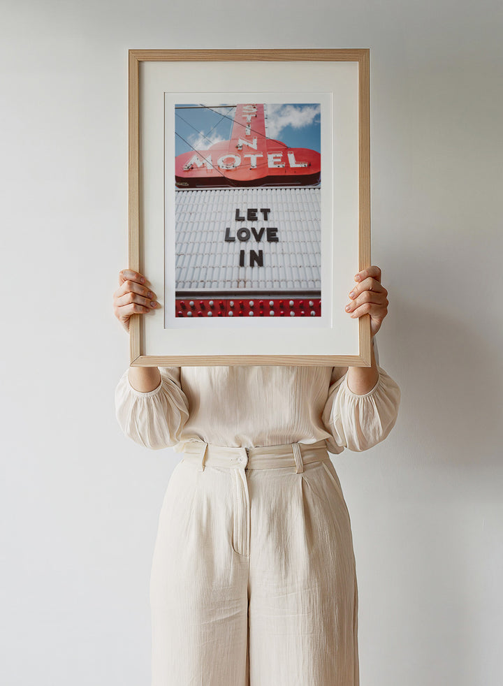 Vintage motel sign with 'Let Love In' message against a blue sky. Let Love In by Bethany Young features a vintage motel sign with red neon lettering against blue sky. Retro Americana photography wall art print.