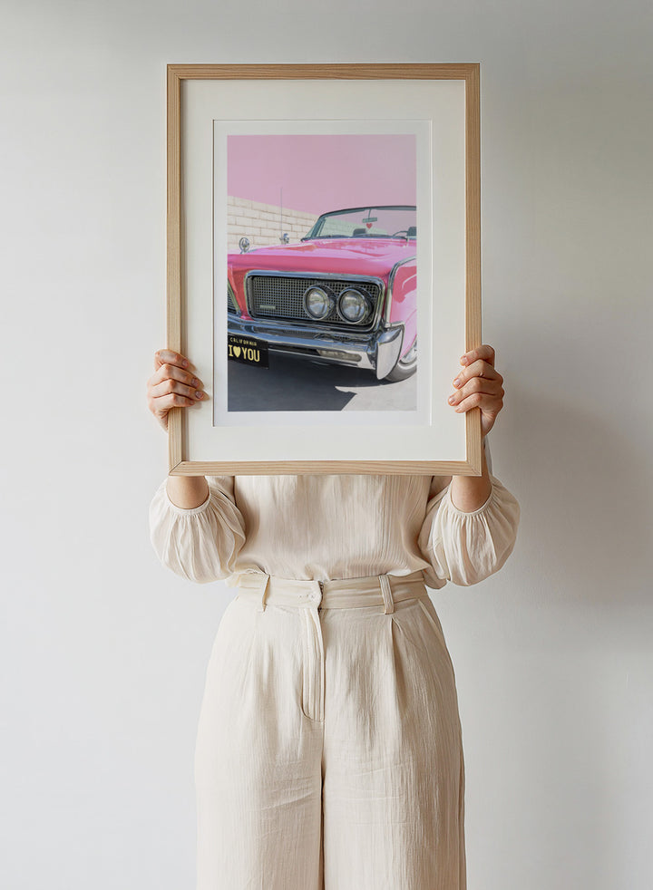 Pink vintage car with 'I ❤️ YOU' license plate against a pink sky. P.S. I Love You by Tom Windeknecht is officially licensed photography featuring a vibrant pop-art style photo of a car with romantic typography. Framed or unframed.