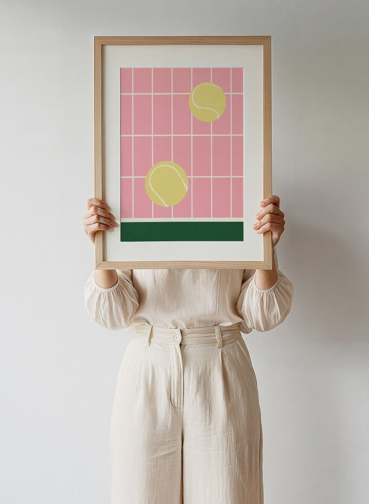 Two yellow tennis balls on a pink grid background with a green stripe at the bottom. Tennis Kitchen by Rosi Feist is a modern minimalist fine art print featuring oversized tennis balls on pink tile. Officially licensed wall poster with optional artisan framing.