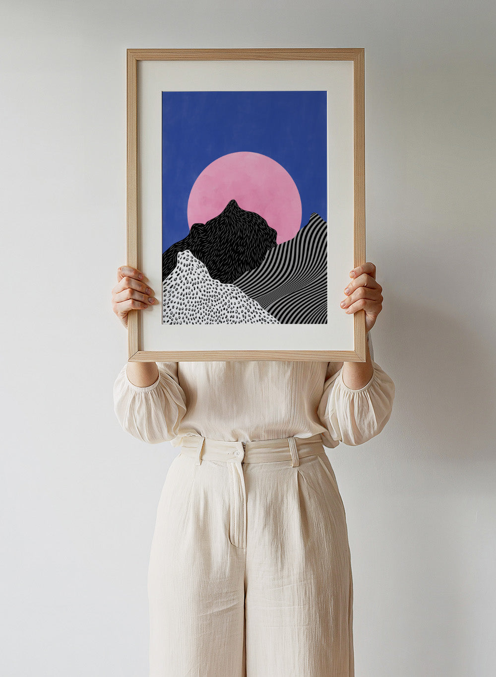 Artistic depiction of mountains with a pink moon in a blue sky. Surreal Peaks View by Elena Ristova is a minimalist abstract landscape print with bold mountains and a pink sun. Officially licensed wall poster with framing.