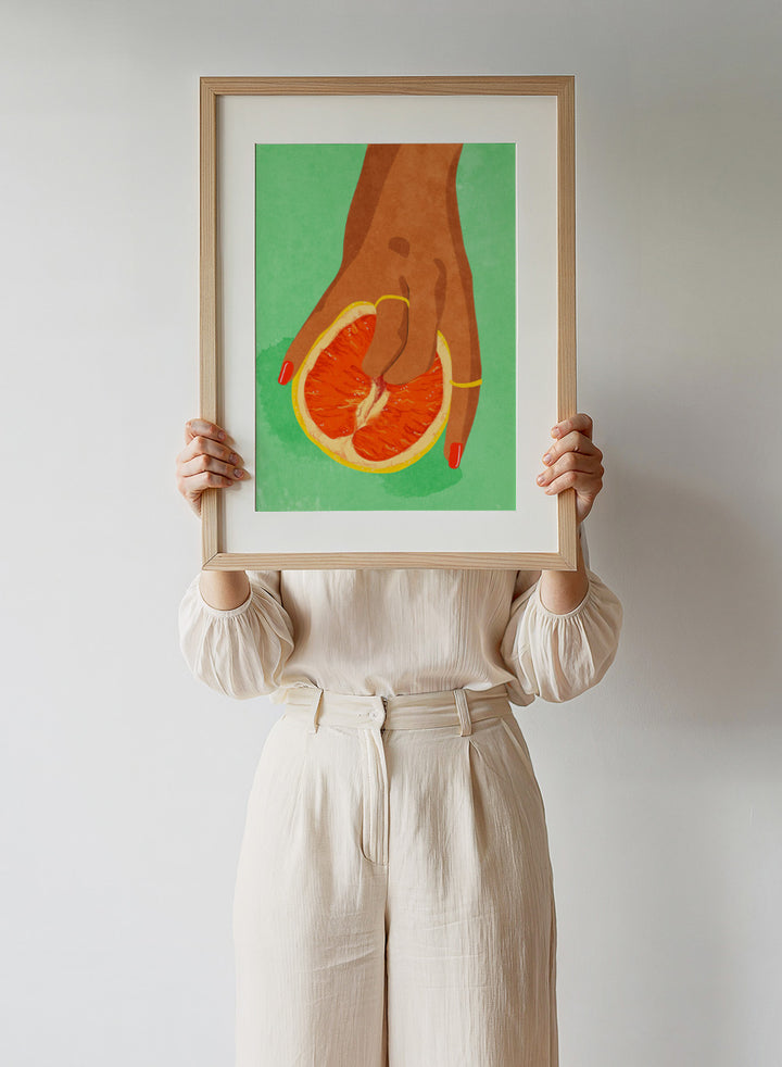 Illustration of a hand holding a sliced orange against a green background. Fruit Lover wall art print by Raissa Oltmanns blends playful citrus and intimate symbolism in a contemporary modern aesthetic poster.