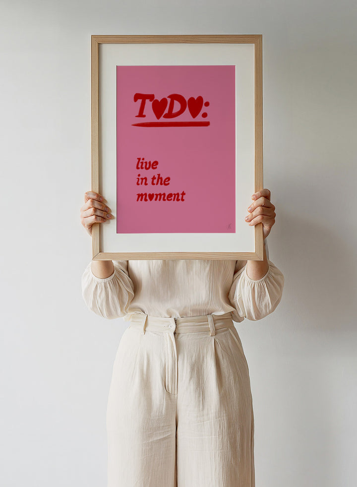 Pink poster with red text 'TODO: live in the moment' on a pink background. Typography wall art reading “live in the moment” with red hearts on pink background by Anne-Marie Volfova. Officially licensed motivational art print.