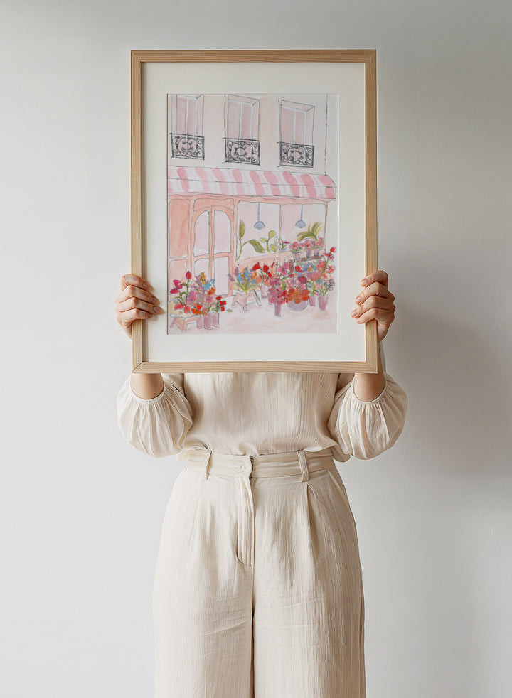 Illustration of a flower shop with a striped awning and flowers in front. Pink Flower Shop by Katharina Puritscher features a Paris-inspired storefront illustration with colorful bouquets, soft watercolor textures, and European street details. Floral art print with optional framing.