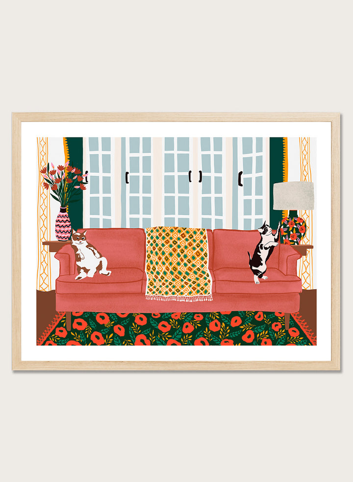 A framed art print depicting a cozy living room scene with a coral-pink sofa, two cats, a patterned throw blanket, and a floral rug. Playful Cats Living Room by Ceyda Alasar cat art print featuring two mischievous cats, a coral sofa, floral rug, patterned throw, and colorful decor.